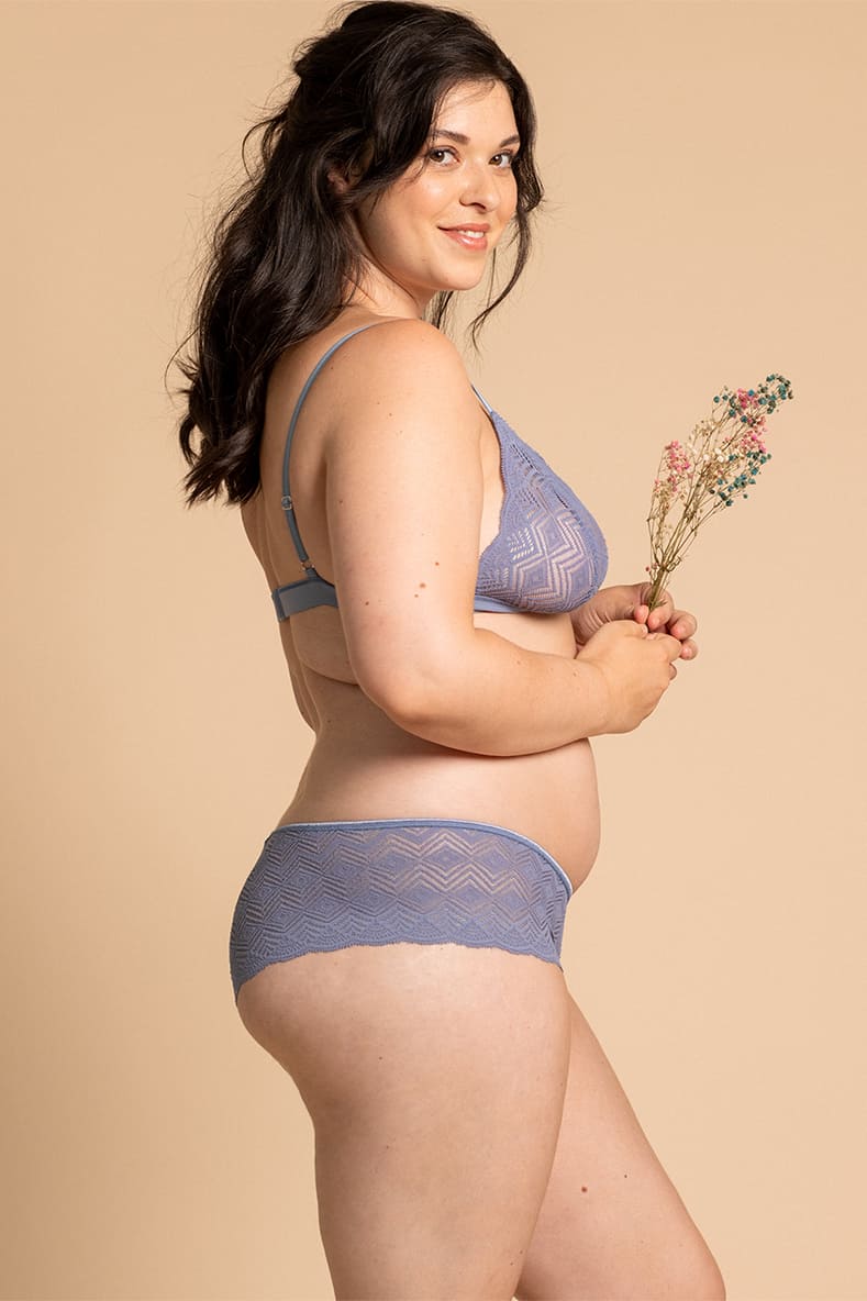 Jeune femme souriante, de profil, en lingerie bleue, elle porte le modèle Dawa myosotis de la marque de lingerie parisienne éco-responsable avec à la main un petit bouquet. 